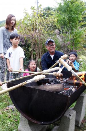 木立の中で棒にパン生地を付けて火で炙る大人と子ども